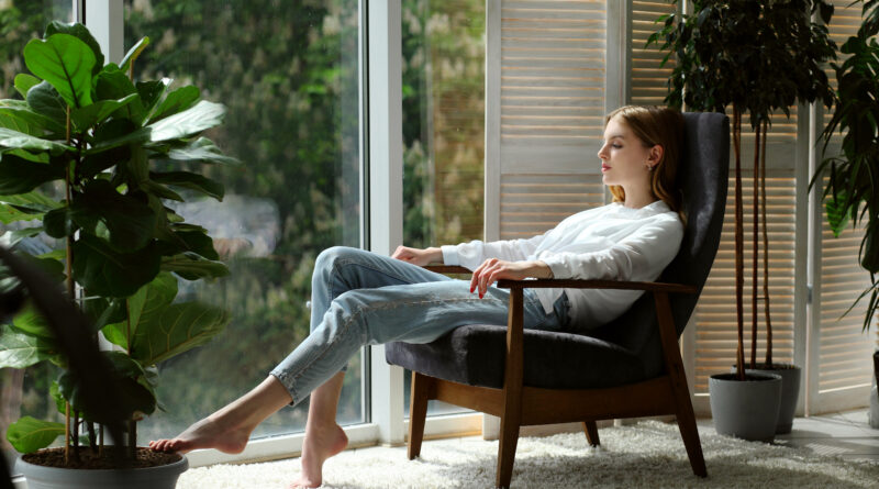 Young woman at home sitting on modern chair near window relaxing in living room